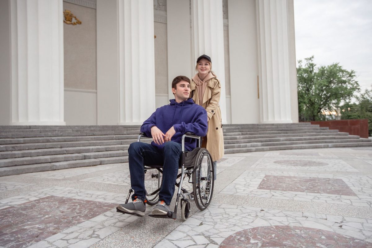 Personne en fauteuil roulant accompagnée devant un bâtiment