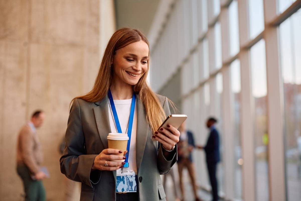 Femme en tailleur consultant son smartphone, un café à la main, dans le hall d'un immeuble de bureaux